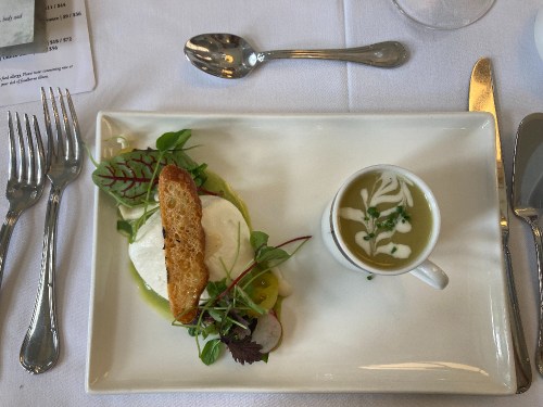 A plate with a poached egg on greens, a slice of toasted bread, and a small cup of soup, set on a white tablecloth.
