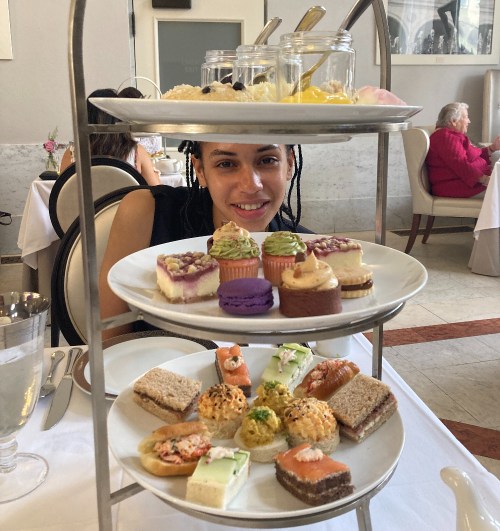 A woman smiles behind a tiered stand filled with colorful pastries and savory snacks at a dining table.