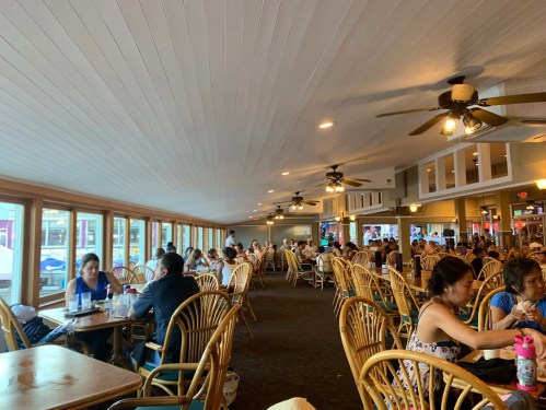 A bustling restaurant interior with wooden chairs, large windows, and diners enjoying their meals.
