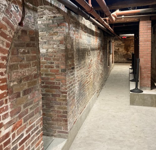 A dimly lit brick corridor with exposed pipes and a sandy floor, featuring archways along the walls.