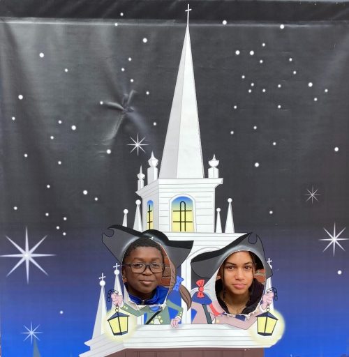 Two people pose in a cutout of a whimsical church with a starry background, wearing hats and smiling.