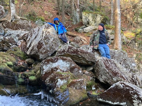 Two people sit on large rocks by a stream in a wooded area, surrounded by trees and mossy stones.