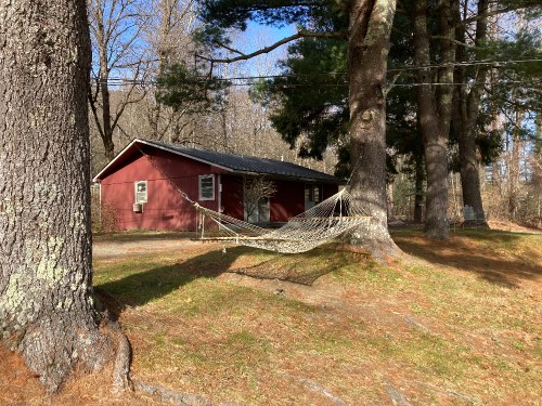 A red cabin surrounded by trees, with a hammock hanging between two large trees in a grassy area.