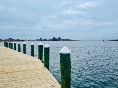A wooden dock extends over calm water, lined with green posts topped with white caps under a cloudy sky.