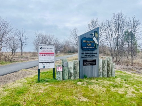 Sign for Ferry Point Park, featuring park rules and directions, surrounded by bare trees and a cloudy sky.