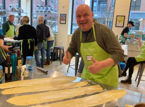 A smiling man in a green apron gives a thumbs up while standing by rolled-out dough in a cooking class.