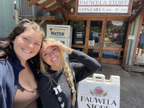 Two young women smile in front of the Pauwela Store, with a sign indicating open hours and a water station nearby.