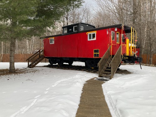 A red caboose on tracks, surrounded by snow and trees, with wooden steps leading up to it.