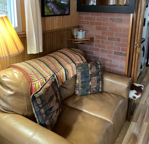 A cozy living room corner featuring a leather sofa with patterned cushions and a colorful throw blanket against a brick wall.