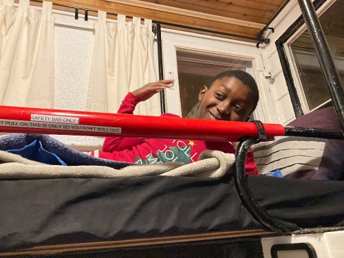 A child smiles while sitting on a bed with a safety bar, surrounded by blankets and a cozy room setting.