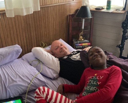 A man and a child relax on a bed, smiling. The child wears festive pajamas, and there are books and a lamp nearby.