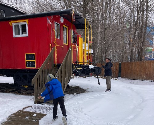 A child in a blue jacket climbs stairs to a red caboose, while an adult stands nearby in a snowy setting.