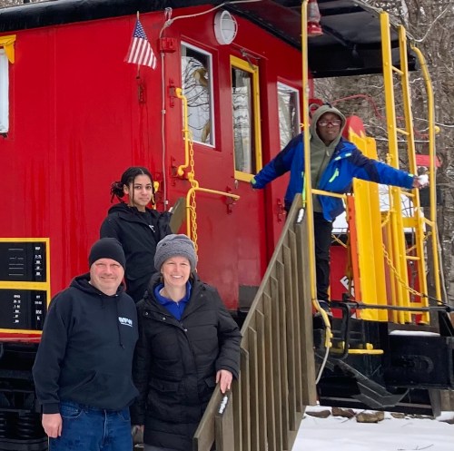 A family poses in front of a red train caboose in winter, with snow on the ground and trees in the background.