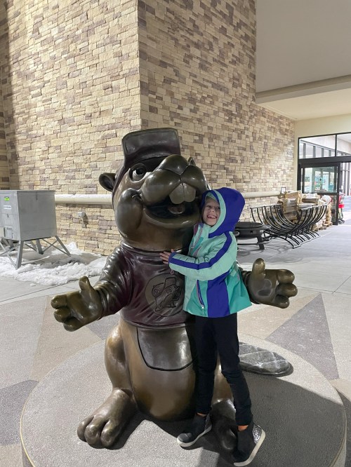 A child in a colorful jacket hugs a large bronze beaver statue in a snowy outdoor setting.