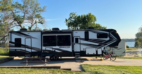 A large, modern RV parked at a campsite near a lake, with a picnic table and a bicycle nearby.