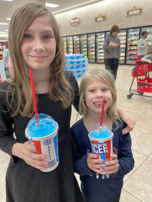 Two young girls smile while holding blue Icee drinks in a store aisle.