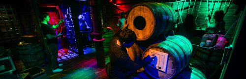 A dimly lit room with colorful lights, featuring barrels and people engaged in an escape room activity.