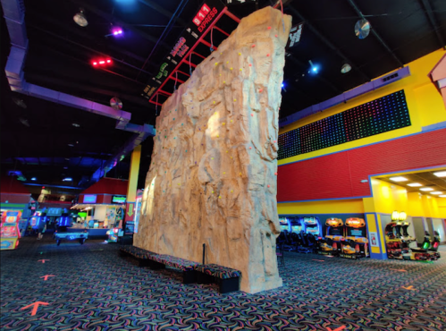Indoor rock climbing wall in a colorful arcade, with arcade games visible in the background and vibrant lighting.