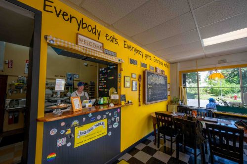 A bright café interior with a welcoming counter, cheerful yellow walls, and a sign promoting community dining.