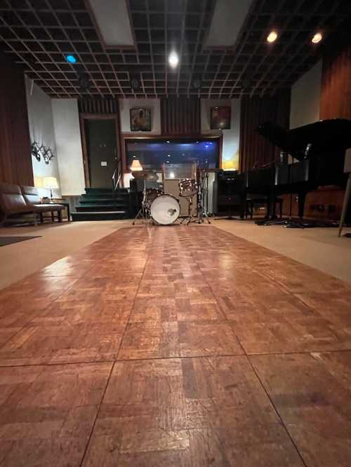 A spacious music studio featuring a drum set, piano, and warm wooden decor, with soft lighting and a cozy atmosphere.