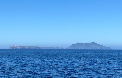 A distant view of two islands against a clear blue sky, with calm ocean waters in the foreground.