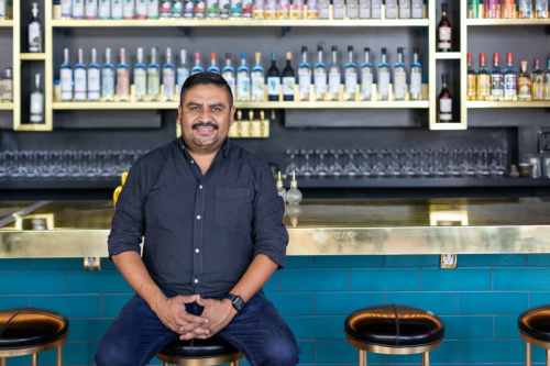 A smiling man sits at a bar with a colorful liquor shelf in the background, showcasing various bottles and glassware.