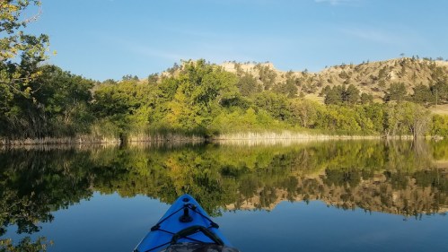 A kayak on calm water, reflecting lush greenery and a hillside under a clear blue sky.