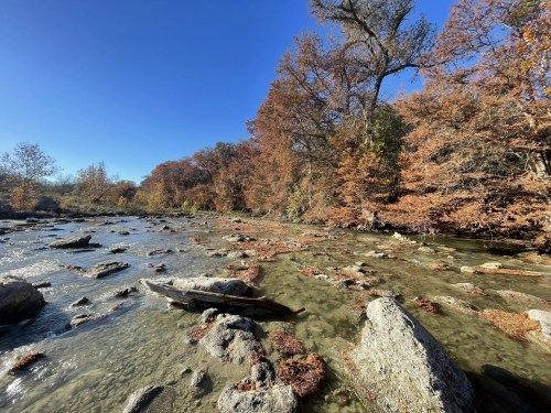 A serene river scene with rocky banks and autumn trees under a clear blue sky.