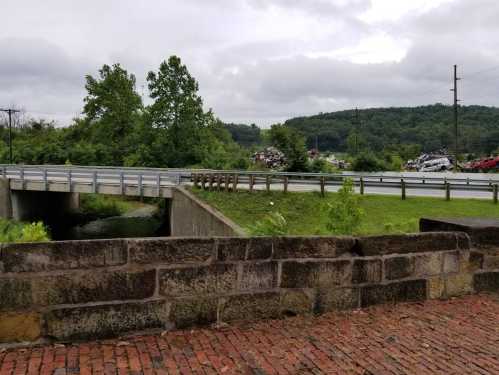 A view of a bridge over a creek, with greenery and a distant junkyard under a cloudy sky.