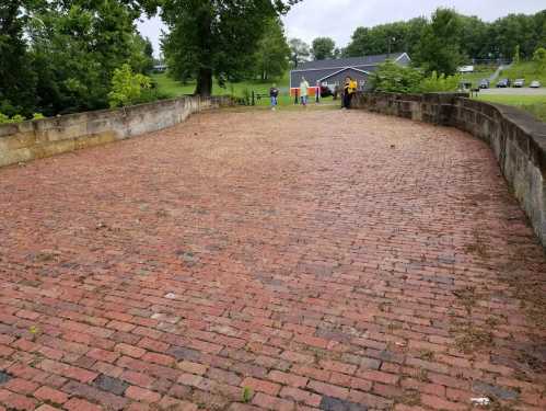 A brick pathway leads to a grassy area with trees, while people stand in the distance near a building.