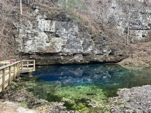 A serene blue spring surrounded by rocky cliffs and trees, with a wooden walkway leading to the water's edge.