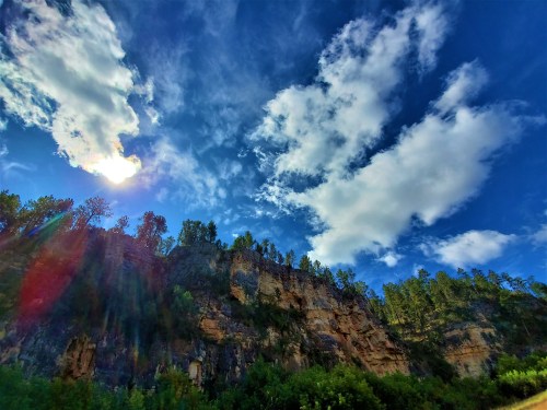 A rocky cliff under a bright blue sky with fluffy clouds and sunlight shining through, surrounded by greenery.
