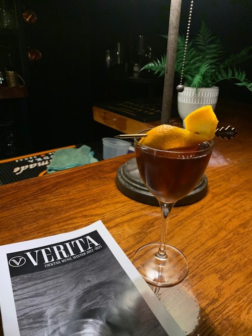 A cocktail garnished with an orange slice and a cherry, placed on a wooden bar next to a menu titled "VERITA."
