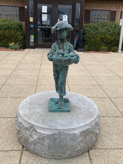 Bronze statue of Johnny Appleseed holding a basket of apples, standing on a stone pedestal outside a building.