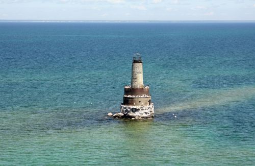 A tall lighthouse stands on a rocky base in the middle of a calm, blue-green sea under a clear sky.
