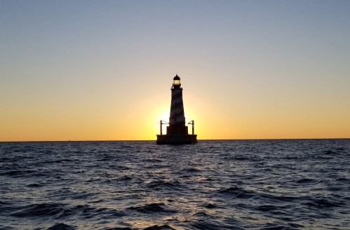 A striped lighthouse stands in the ocean at sunset, with the sun glowing behind it, casting a warm light.