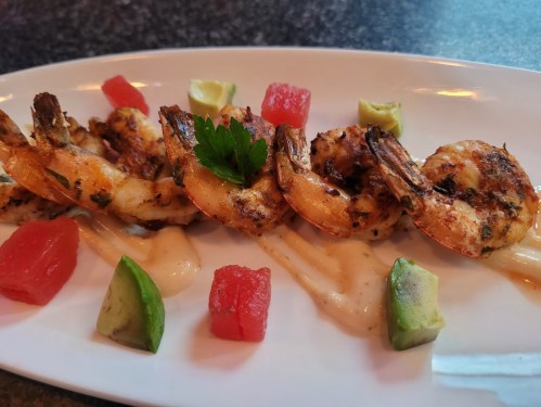 Grilled shrimp on a plate, garnished with avocado, watermelon cubes, and a drizzle of sauce.