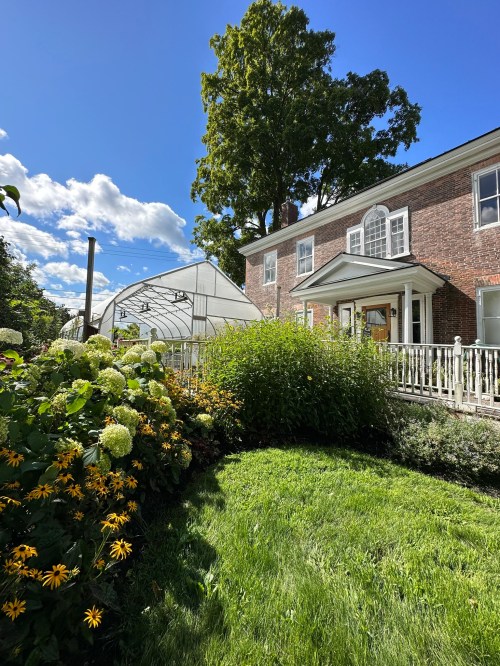 A brick house with a greenhouse, surrounded by vibrant flowers and lush greenery under a blue sky with clouds.