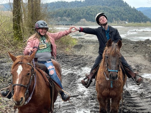 Two riders on horseback make a heart shape with their hands, smiling against a scenic backdrop of trees and mountains.