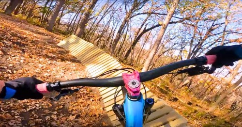 A mountain biker's view riding on a wooden ramp through a forest with autumn leaves.