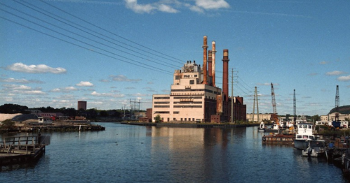 A large industrial building with smokestacks beside a calm river, surrounded by boats and power lines under a blue sky.