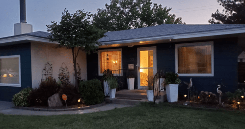 A cozy house with a blue exterior, warm lights, and a well-kept garden, set in the evening.
