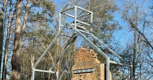 A metal frame structure surrounds the ruins of a stone building, set among trees under a clear blue sky.