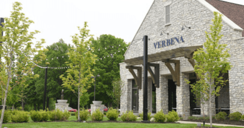 A stone building with "VERBENA" signage, surrounded by green trees and landscaping on a cloudy day.