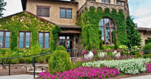 A charming stone building covered in greenery, surrounded by colorful flower beds and a landscaped pathway.