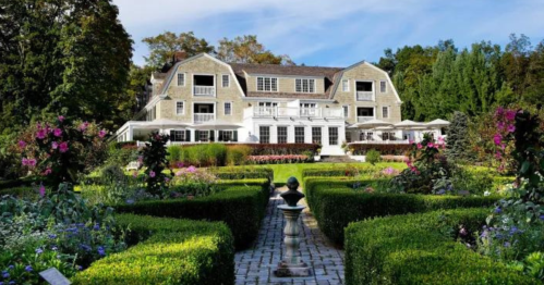A large, elegant house surrounded by lush gardens and neatly trimmed hedges under a clear blue sky.