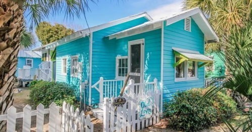 A charming blue cottage with white trim, surrounded by greenery and a picket fence, under a clear blue sky.