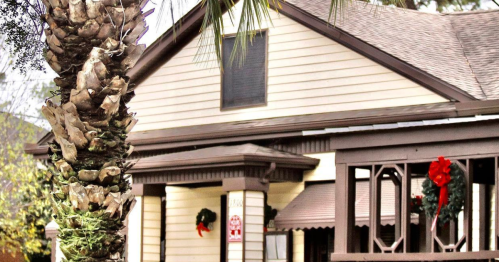 A cozy house decorated for the holidays, featuring wreaths and a red bow, with a palm tree in the foreground.