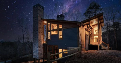 A modern house at night, illuminated against a starry sky, surrounded by trees.