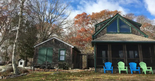 A cozy cabin with a screened porch, surrounded by trees and colorful autumn leaves, featuring blue chairs in front.